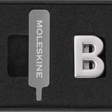 Sleek silver Moleskine Pins B for personalizing notebooks with initials or phrases, crafted from durable zinc alloy.