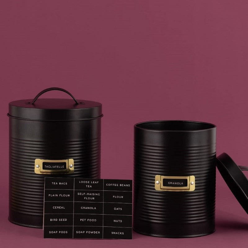 Black storage canister set featuring a ribbed design, gold window frame, and eco-friendly steel; includes two sizes and labels.