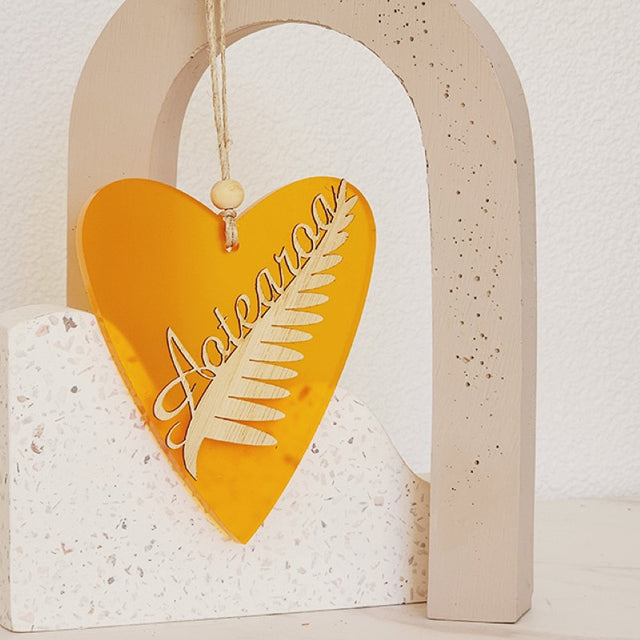 Heart-shaped hanging ornament crafted from orange satin acrylic, featuring intricate silver fern designs, ready to hang.